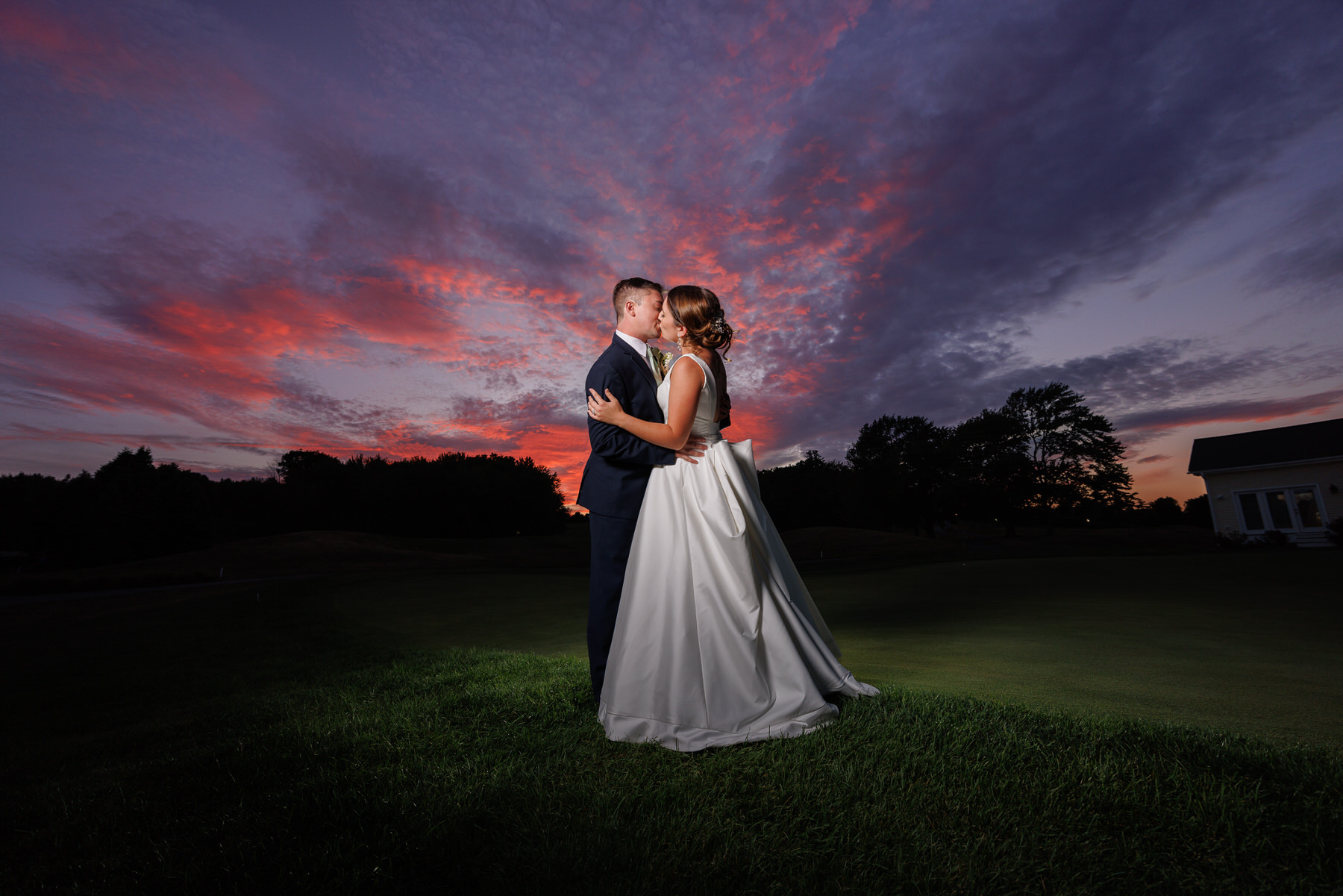 Caroline and Eric-720-1 A couple kisses on a golf course at sunset, with a vibrant pink and blue sky above and a white building in the background, perfectly capturing their wedding timeline.