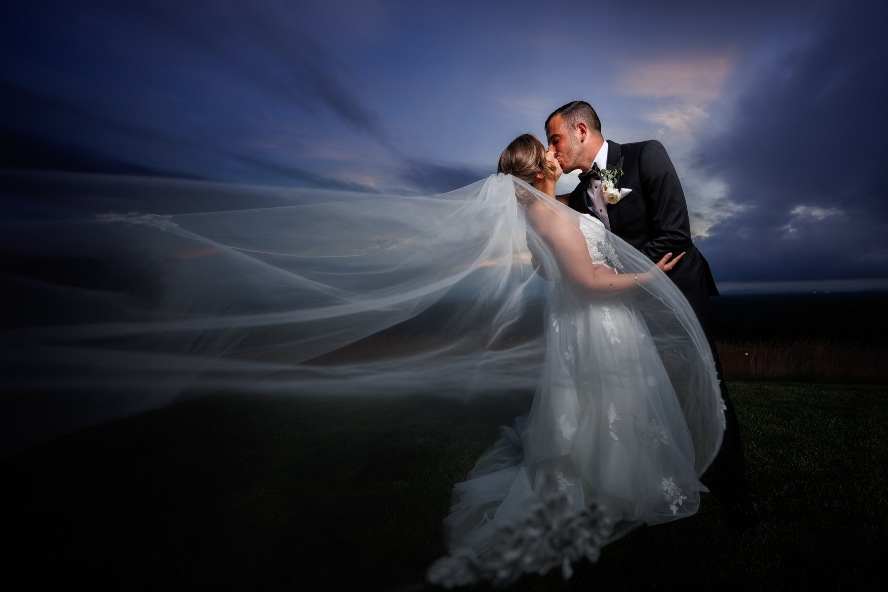 GM4_7367-1 Bride and groom kissing outdoors at dusk, with the bride's long veil elegantly blowing in the wind under a dramatic cloudy sky, captured perfectly in their wedding photography.