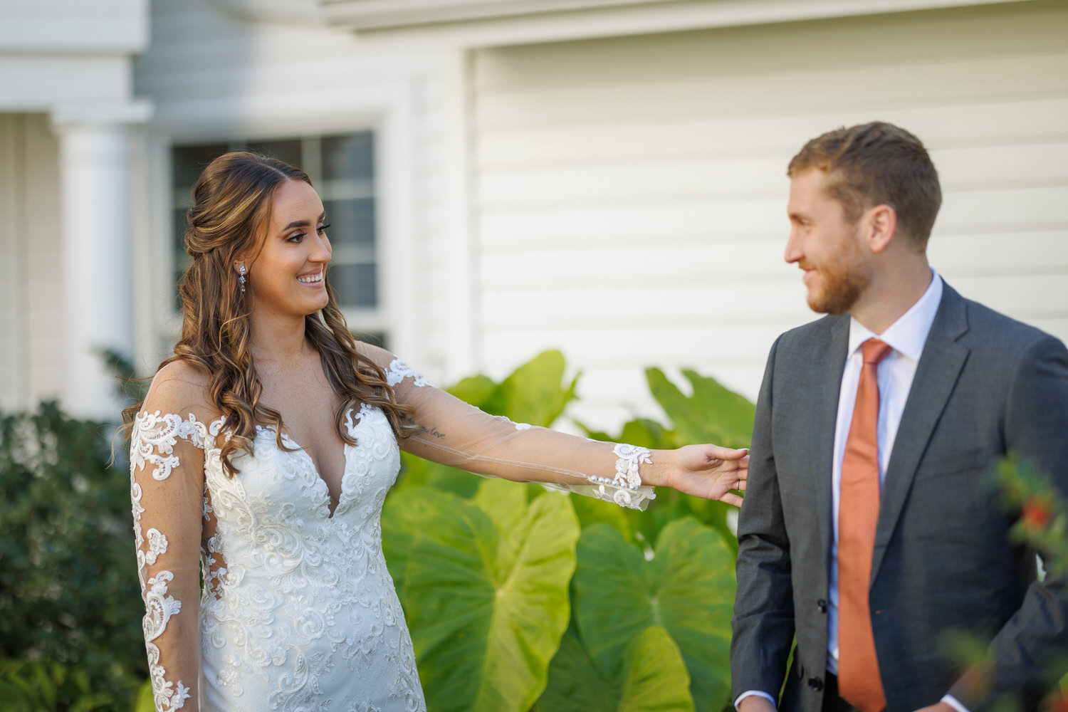 Kelly and Joe-1010-1 Bride in a lace dress smiling at the groom in a suit, pinning a flower on his lapel outdoors during their wedding photography session, with a white house in the background.