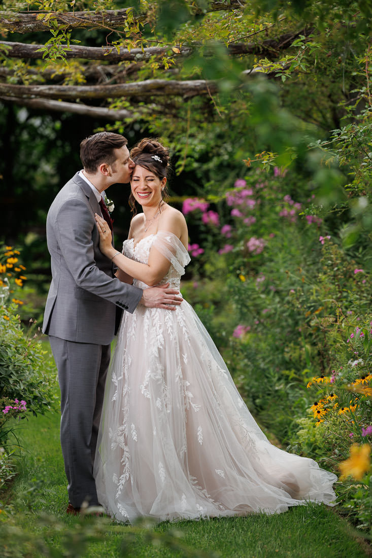 Union Station Wedding-12 A bride in a white floral dress and a groom in a gray suit kissing in a lush garden surrounded by vibrant flowers at their Northampton Ma wedding.