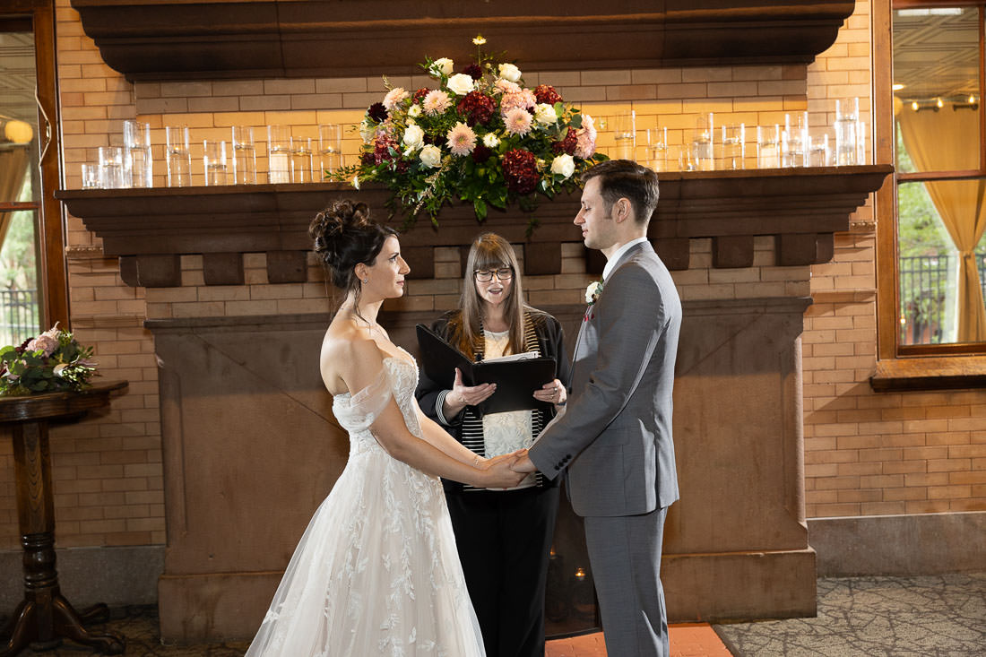 Union Station Wedding-13 Briana and Jacob, holding hands at the altar with an officiant between them, in a venue decorated with flowers and candles at Union Station Wedding in Northampton, MA.