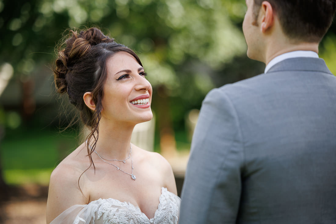 Union Station Wedding-6 A bride, Briana, smiling joyfully at the groom, Jacob, in an outdoor setting in Northampton, MA. She is wearing a white dress with lace details and a necklace.
