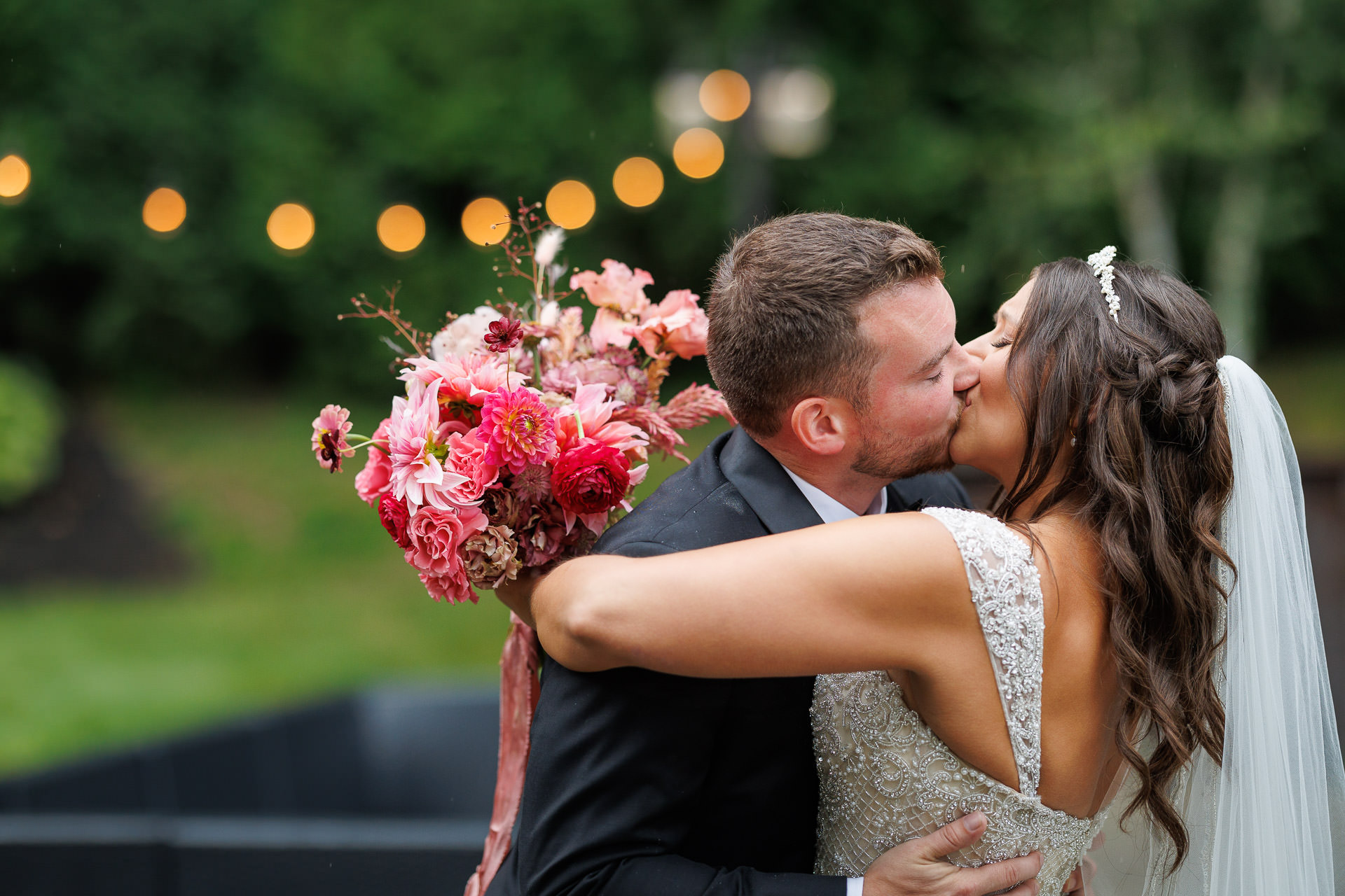 A bride and groom kissing outdoors at the Mountain Rose Inn while holding a bouquet of pink flowers, with blurred lights in the background.