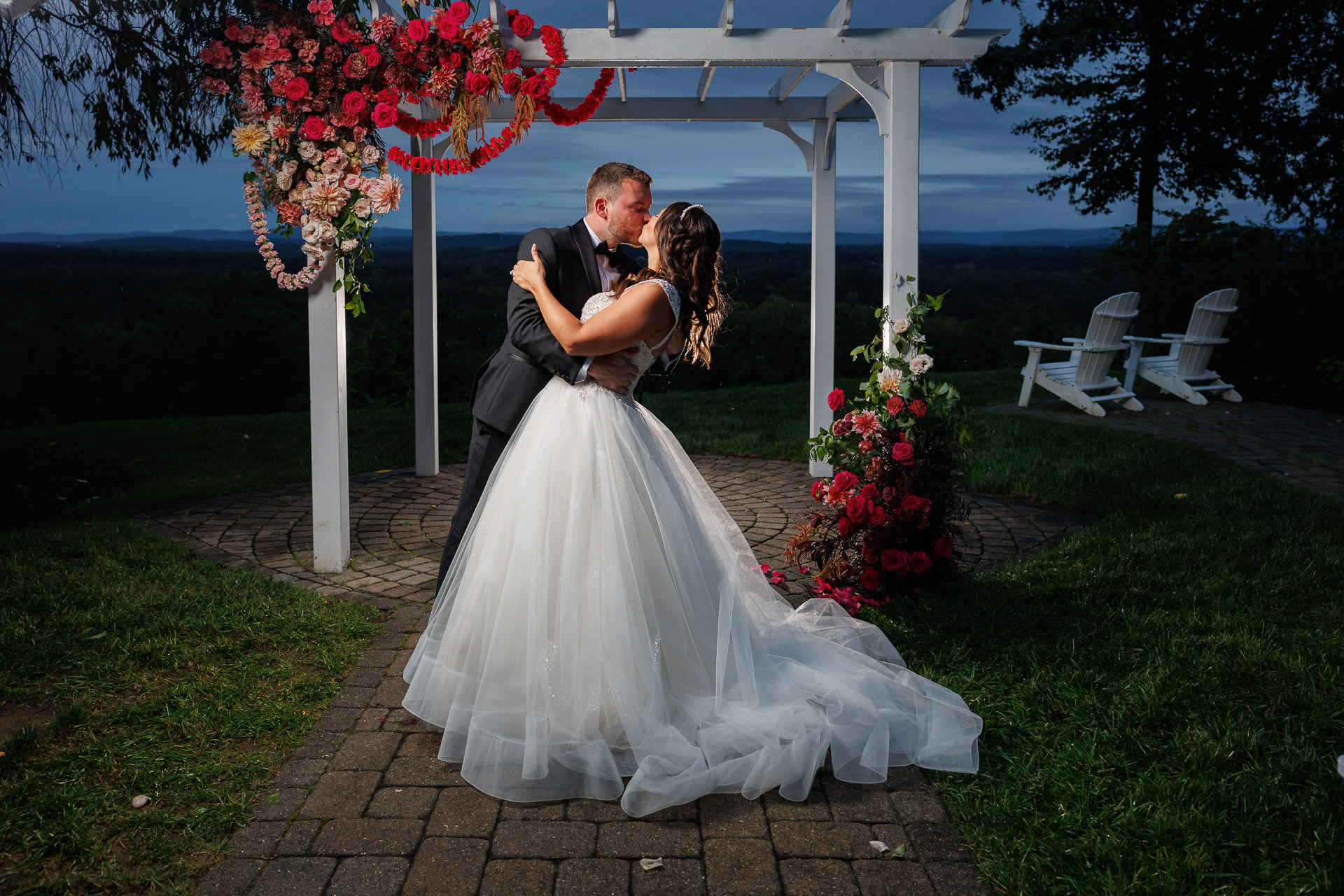 A bride and groom embrace under a gazebo adorned with colorful flowers at the Mountain Rose Inn, with a scenic dusk sky in the background.