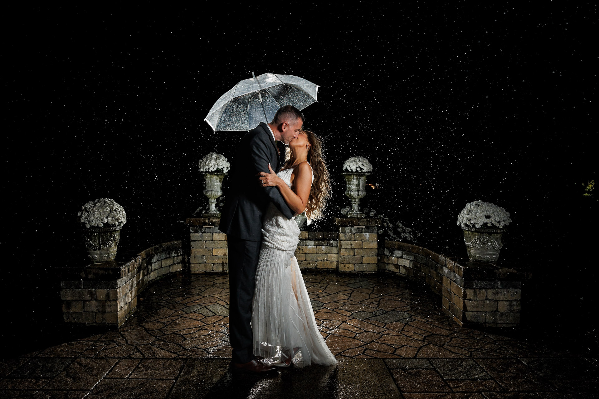 Erica and Jeff kiss under a transparent umbrella at night, with rain visible in the light against a dark, patterned ground and illuminated floral pedestals on either side.