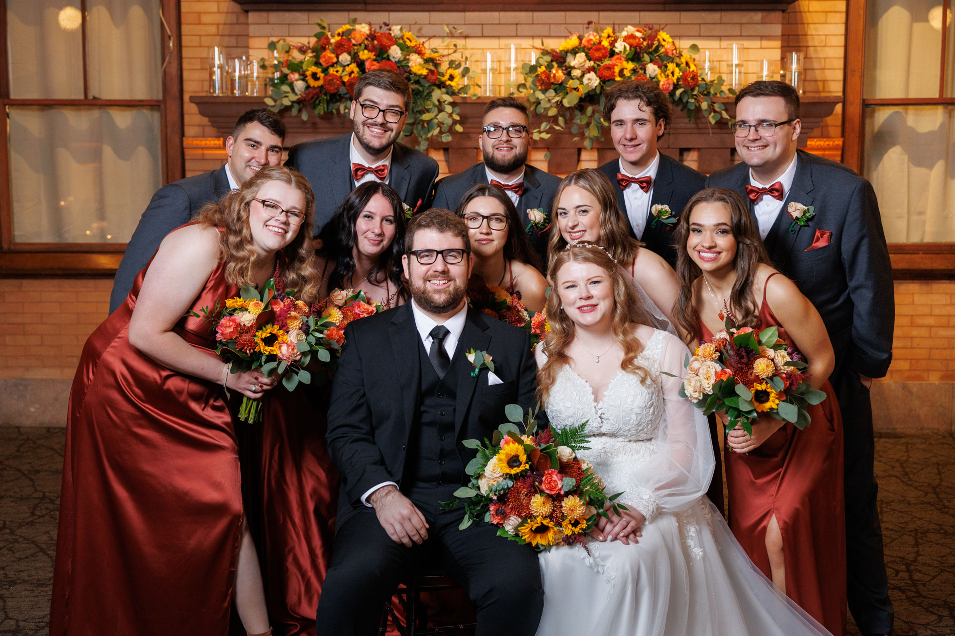 Bridal couple surrounded by bridesmaids and groomsmen in red dresses and suits, posing with bouquets in front of a floral backdrop at Union Station Wedding.