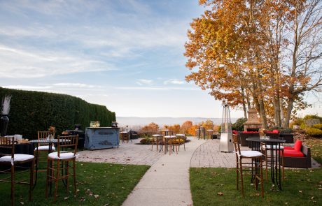 Outdoor patio with scattered seating, high-top tables, and trees with autumn foliage, set against a backdrop of mountains under a blue sky.