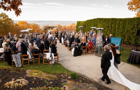 A bride and groom walk down the aisle during an outdoor wedding ceremony attended by seated guests. The setting features autumn foliage, heaters, and a scenic backdrop of hills and sky.