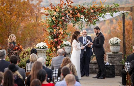 A couple exchanges vows under a floral arch during an outdoor wedding ceremony, with a woman officiant and guests seated in the foreground. The scene features autumn foliage and decorative flowers.