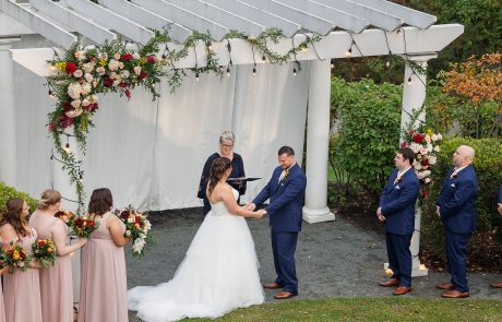 Caroline and Matt-341 A couple stands, holding hands, in an outdoor wedding ceremony under a decorated pergola. A celebrant officiates, while bridesmaids in pink and groomsmen in blue look on.