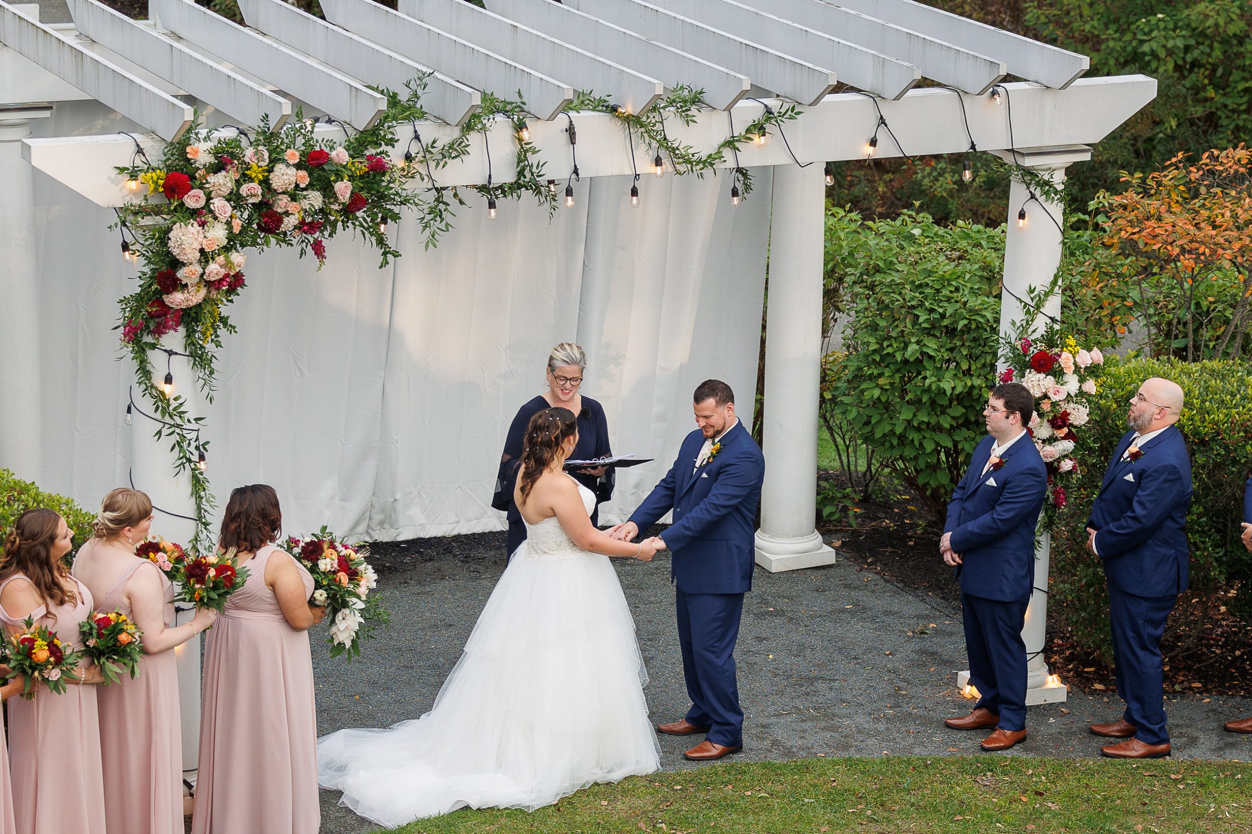 Caroline and Matt-341 A couple stands under a pergola exchanging vows during their outdoor wedding ceremony. Bridesmaids in pink dresses and groomsmen in blue suits stand on either side. A greenery and floral arrangement adorns the pergola. The Inn on Boltwood wedding