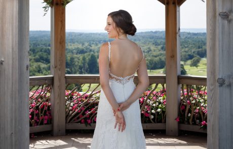 A bride in a white sleeveless wedding gown stands with her back to the camera under a wooden arch adorned with flowers at The Starting Gate Wedding venue, overlooking a scenic landscape.