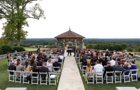The Starting Gate Wedding features an outdoor ceremony with guests seated on either side of an aisle leading to a charming wooden gazebo, set against a scenic landscape of trees and distant hills.