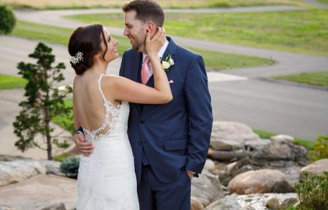 A bride in a white dress holds the face of a groom in a blue suit as they pose on a rocky path at The Starting Gate Wedding, with greenery and a road in the background.