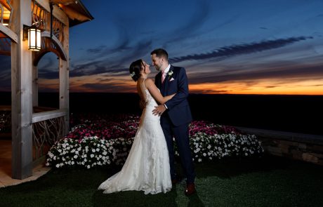 A bride and groom stand smiling at each other under a wooden structure with a sunset in the background. The bride wears a white gown and the groom is in a navy suit. They are surrounded by flowers, creating the perfect scene for their Starting Gate Wedding.