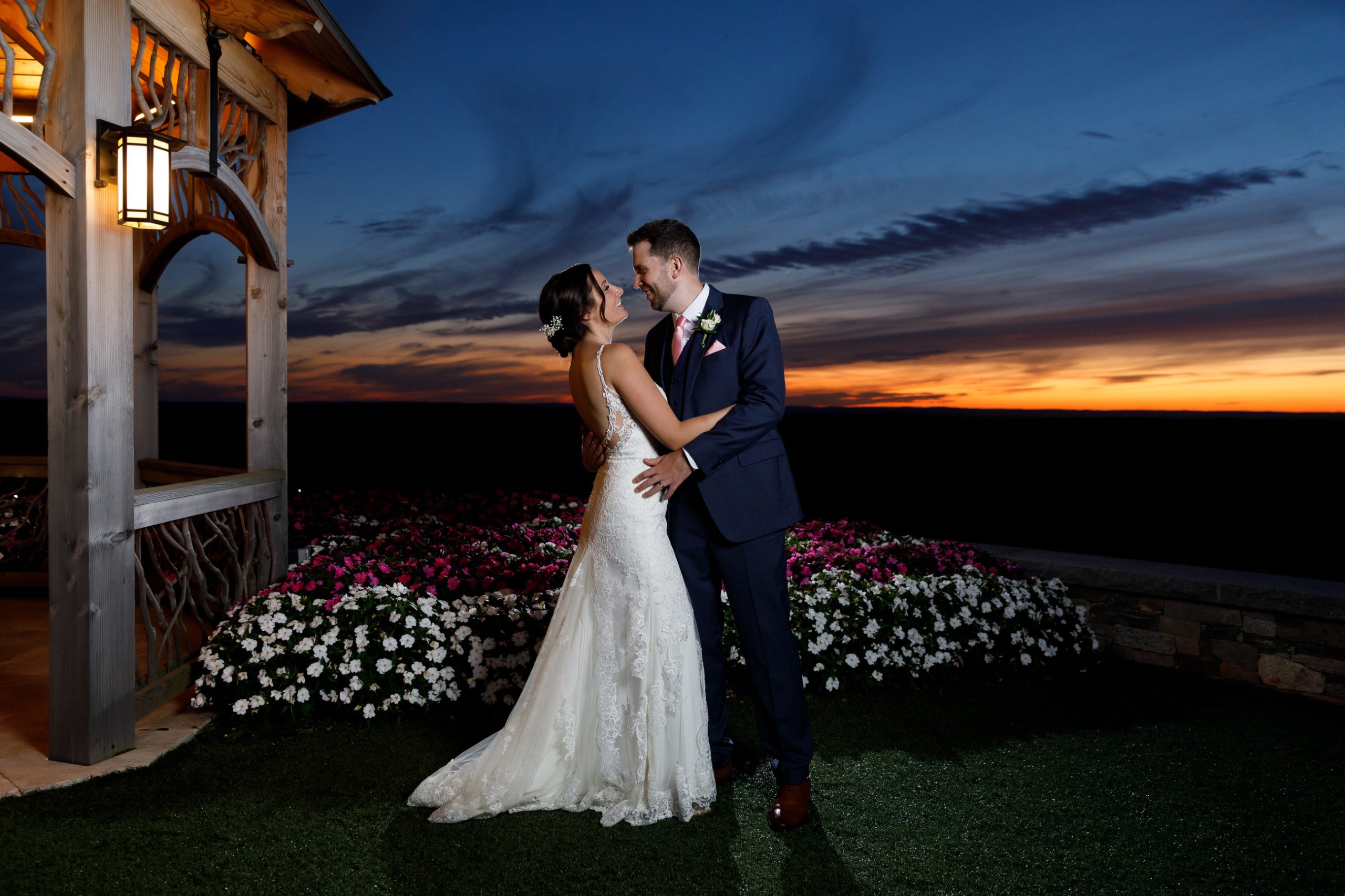 A couple in wedding attire stands outdoors at sunset, under a wooden structure with lantern, surrounded by white and pink flowers at The Starting Gate Wedding.