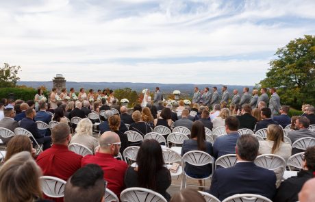 An outdoor wedding ceremony with a couple getting married. The seated guests are watching, many wearing formal attire. Bridesmaids and groomsmen stand on either side of the couple.