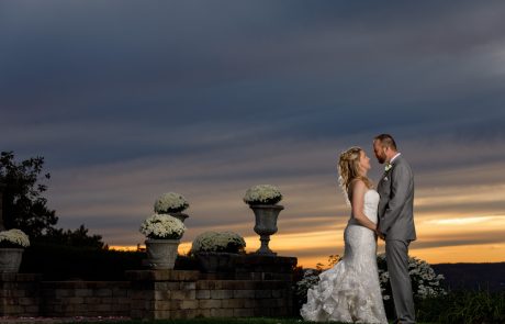 A bride and groom stand facing each other, holding hands, against a sunset sky. White flowers in stone planters are visible in the background.