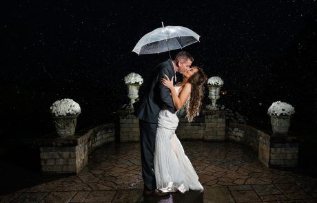 A couple in wedding attire kisses under an umbrella in the rain at night, standing on a stone patio with large flower arrangements on pedestals in the background.