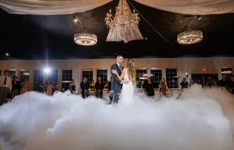 A bride and groom share a first dance surrounded by fog, under chandeliers in an elegantly decorated venue, with guests watching from the background.