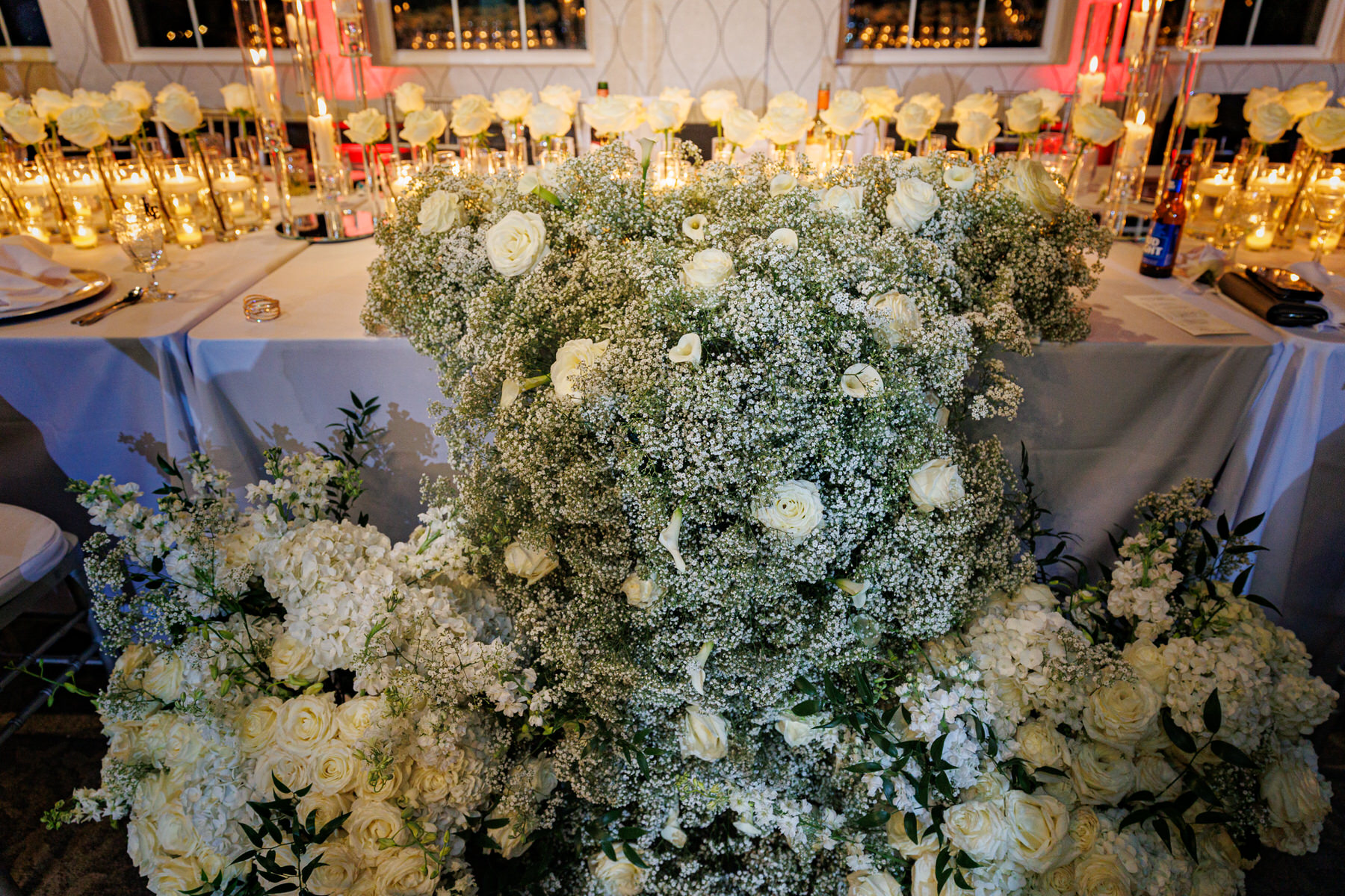 A lavish floral arrangement of white roses, baby's breath, and other white flowers, displayed in front of a decorated table with multiple candles.