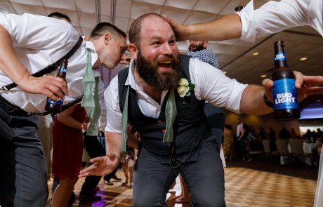 Man in a vest and tie with a boutonniere celebrates at an indoor event, holding a Bud Light bottle while others interact with him energetically. The Log Cabin Weddings