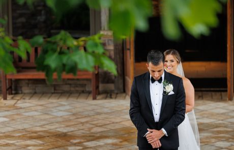 A bride in a white dress and veil stands behind a groom in a black tuxedo, both under a tree with green leaves on a paved area at The Starting Gate Wedding venue. The groom faces forward while the bride looks at him, smiling.