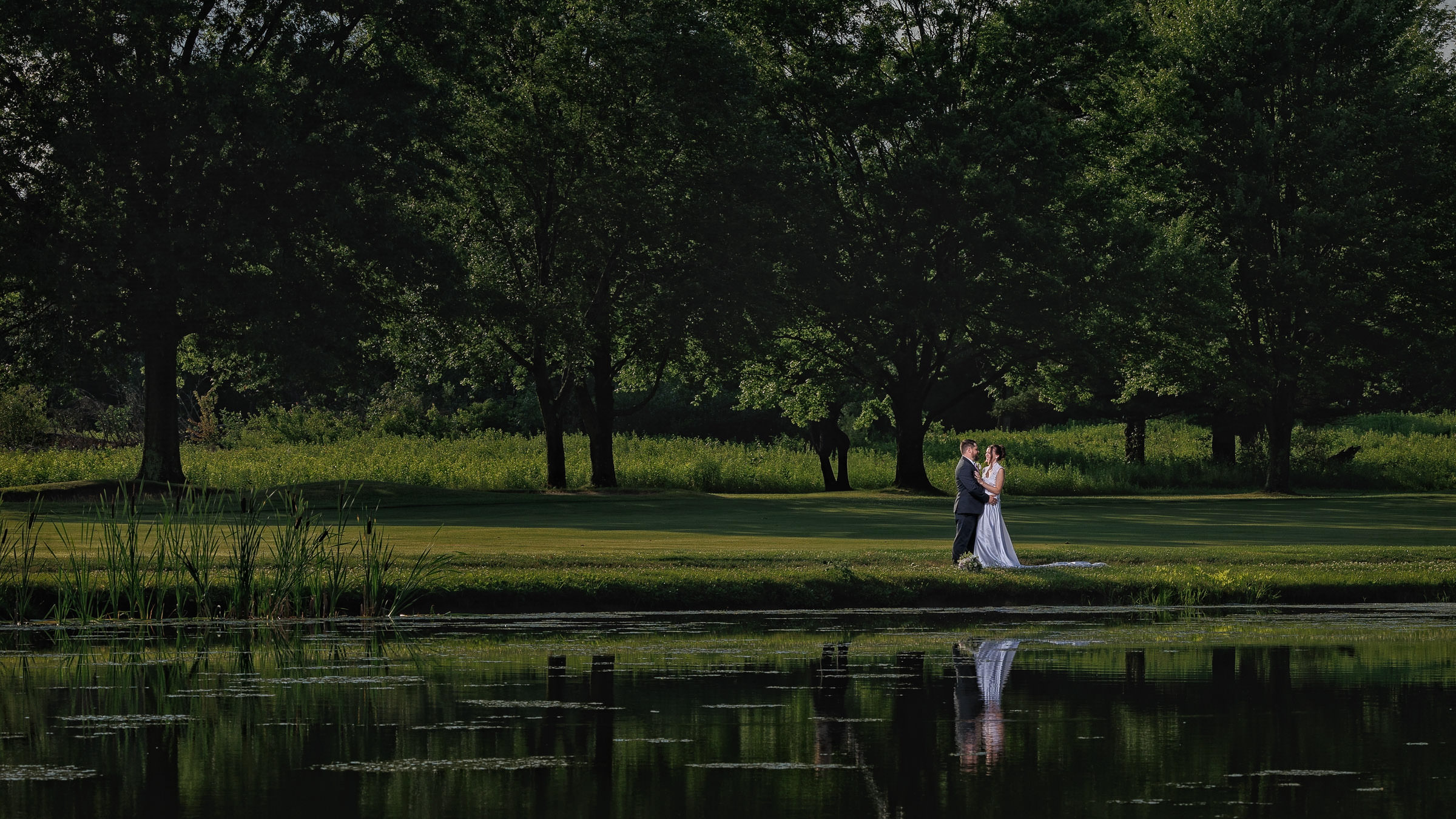 A couple in wedding attire embraces on a grassy area by a pond at Crestview Country Club, surrounded by trees, with their reflection visible in the water.