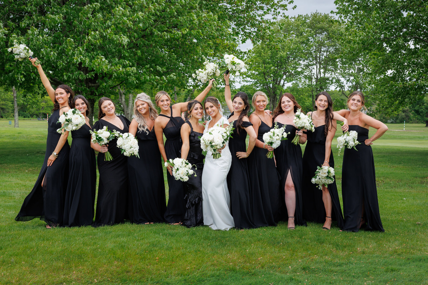 A group of women in black dresses and one in white stand outdoors on grass, holding white floral bouquets, with trees in the background.Bridal party at the Springfield. CC