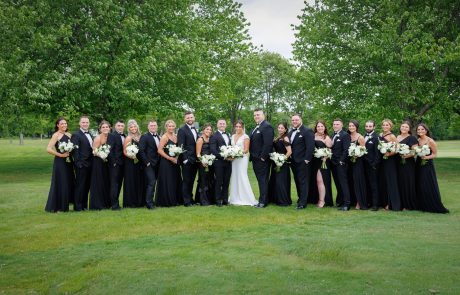 A wedding party stands outdoors, posing for a group photo. The bride and groom are in the center, surrounded by bridesmaids in black dresses and groomsmen in black suits. Everyone is holding white bouquets.
