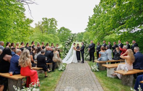 A wedding ceremony taking place outdoors, with the couple standing at the altar surrounded by greenery. Guests are seated on wooden benches, forming two rows on either side of the aisle.