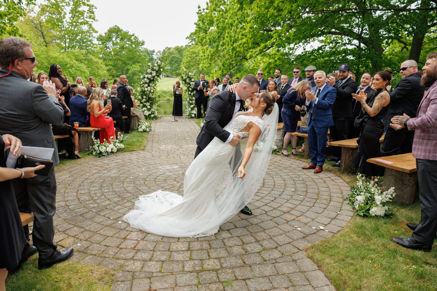A bride and groom share a kiss at their outdoor wedding ceremony, surrounded by guests who are clapping and smiling. The couple stands on a paved circular area amidst lush greenery.