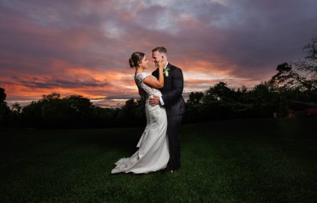 A couple in wedding attire shares an intimate moment on a grassy field at sunset, with a colorful sky as the backdrop. Springfield Country Club Weddings
