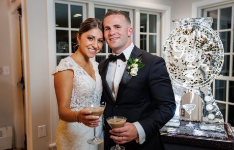A bride and groom, both dressed formally, stand close together holding drinks. They are smiling and posing in front of a large decorative ice sculpture in a well-lit room.
