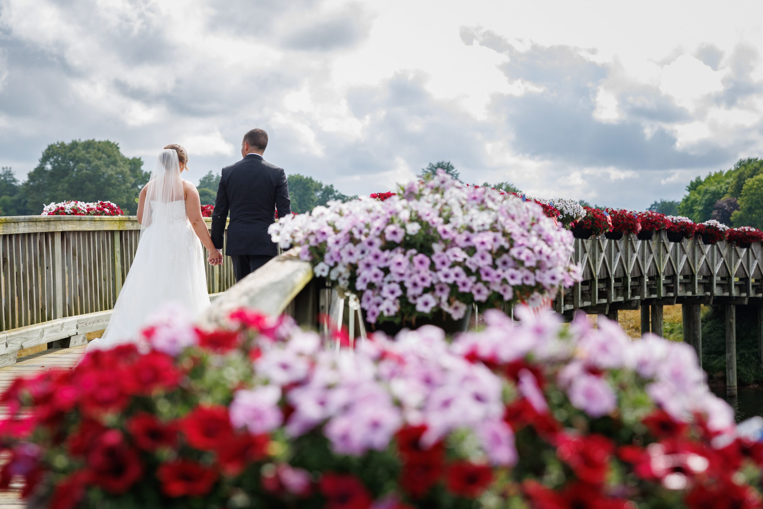 A bride and groom holding hands walk along a wooden bridge adorned with red and white flowers under a cloudy sky, capturing their first steps together at The Starting Gate Wedding.
