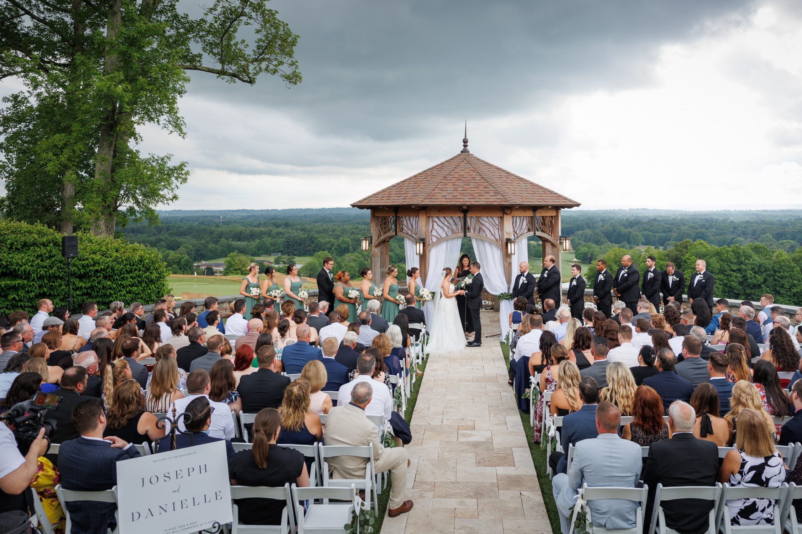 The Starting Gate Wedding ceremony takes place outdoors with a large audience seated, facing a small gazebo where the couple, officiant, and wedding party stand. The background features a scenic landscape under a cloudy sky.