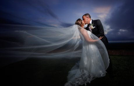 A bride and groom share a kiss outdoors at dusk at The Starting Gate Wedding venue. The bride's veil flows dramatically in the wind, and both are dressed in wedding attire. The sky is darkening, creating a romantic atmosphere.