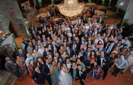 A large group of people dressed formally, gathered in a ballroom under a chandelier, posing for a photo with the bride and groom at the center. This memorable moment at The Starting Gate Wedding captures the joy and elegance of their special day.