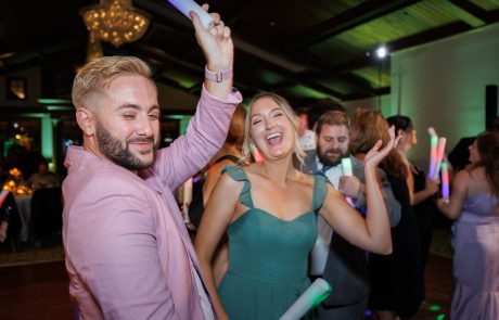 People dancing and smiling at The Starting Gate Wedding, illuminated by colorful lights and holding light sticks. A man in a pink jacket and a woman in a green dress are in the foreground, enjoying the moment.