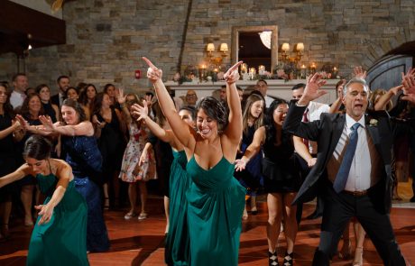 People in formal attire are dancing energetically at The Starting Gate Wedding celebration, with a stone wall and lit candles in the background.