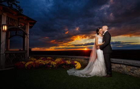 A bride and groom pose together in wedding attire at sunset at The Starting Gate Wedding venue, with a lit wooden structure on the left and colorful flowers in the foreground. Dark clouds fill the sky, creating a dramatic background.