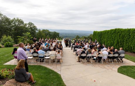 An outdoor wedding ceremony with guests seated on folding chairs facing the couple and officiant at the altar, set against a scenic backdrop of trees and hills. The Log Cabin Weddings