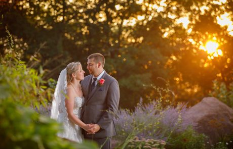 A bride and groom stand holding hands, gazing at each other in an outdoor setting with greenery and a glowing sunset in the background.