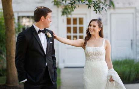Kaitlin and Dan-166 A bride in a white dress smiles and touches the shoulder of a groom in a black suit outdoors in front of a white building.