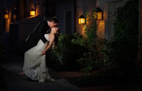 Kaitlin and Dan-686 A couple dressed in formal attire shares a romantic dance pose at night, illuminated by warm wall lights in a dimly lit outdoor setting. The woman in a white gown is being gently dipped by the man.