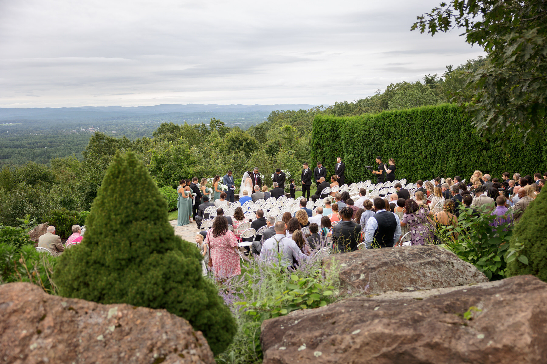 A wedding ceremony is taking place outdoors with mountains in the background. Guests are seated in rows of white chairs facing a couple being married. The event is surrounded by greenery and trees.