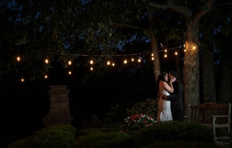 A bride and groom kiss under string lights during an outdoor evening wedding. Trees and a bench are in the background.