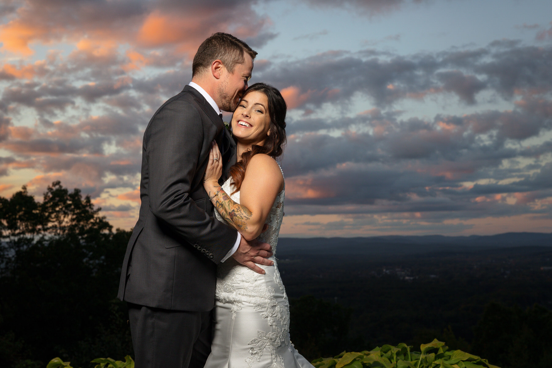 A couple in formal attire embraces and smiles outdoors at sunset, with a view of a scenic landscape and cloudy sky in the background.. The Log Cabin Weddings