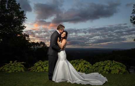 A newlywed couple stands lovingly on a grassy hill at sunset, with a picturesque landscape and dramatic cloudy sky in the background. The bride is in a white gown, and the groom is in a dark suit.