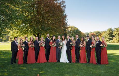 A bride and groom stand outdoors with their wedding party, dressed in red gowns and black suits, arranged in a line in front of trees and greenery.